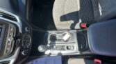 Inside a car cabin focusing on the center console: touchscreen, climate controls, gear selector, and cupholders near the patterned seat upholstery.