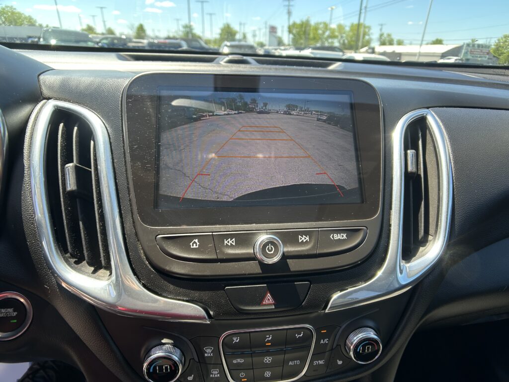 Infotainment screen showing a car’s rear‑view camera, with parking guidelines over a busy lot and cars in the distance.