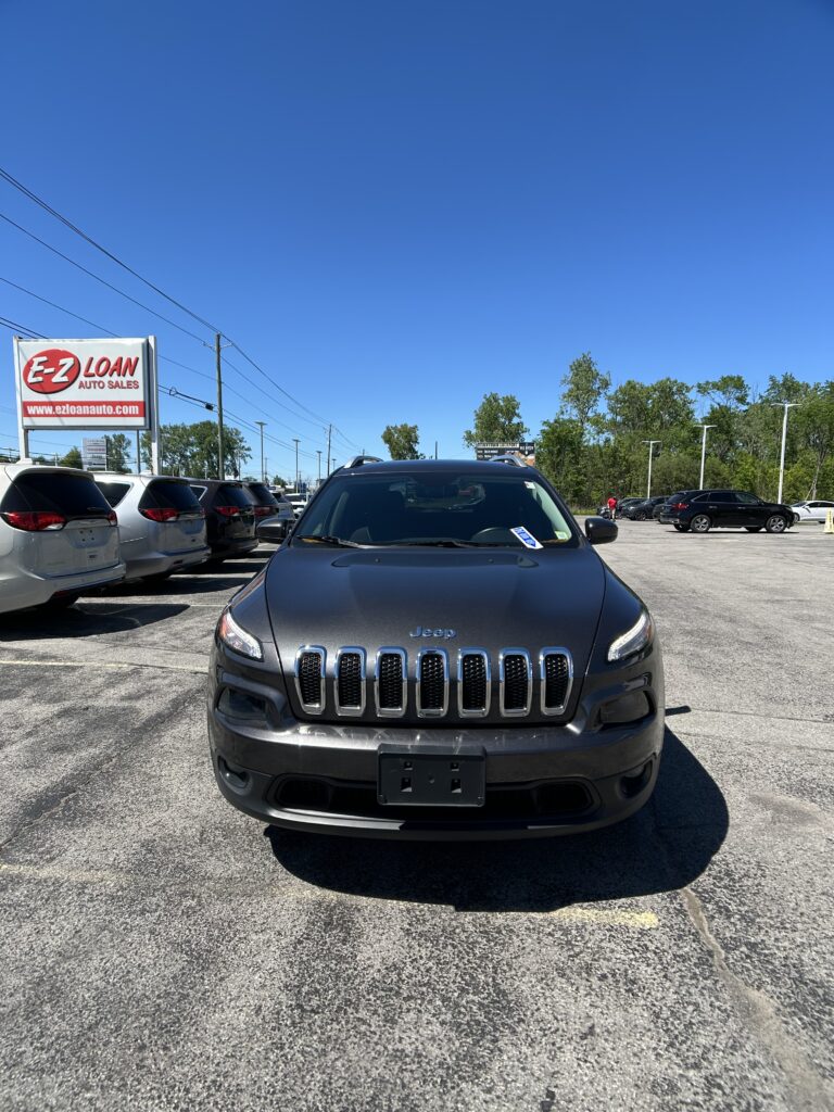 Front view of a dark gray Jeep SUV parked in a car dealership lot, with an E-Z LOAN Auto Sales sign on the left and a blue sky above.