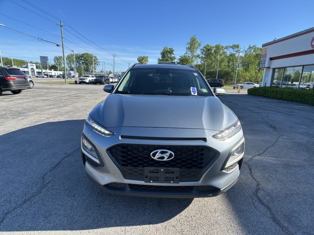 Front view of a silver Hyundai SUV parked in a car dealership lot with a showroom building in the background under a blue sky.