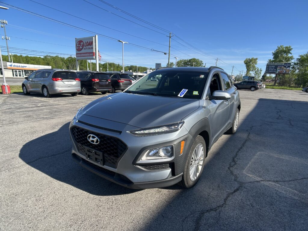 Front-right view of a gray Hyundai SUV in a car dealership lot, with other vehicles and an EZ LOAN Auto Sales sign in the background on a bright day