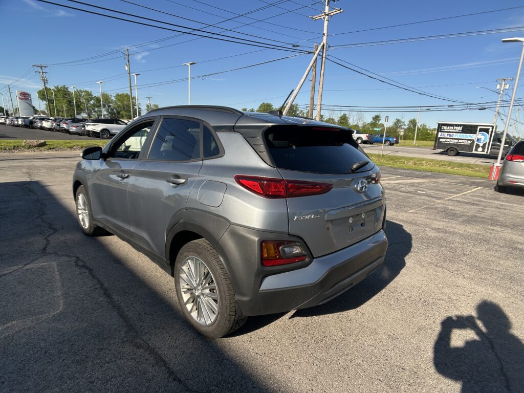 Rear three-quarter view of a gray Hyundai Kona SUV in a car dealership lot with power lines overhead and a clear blue sky