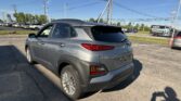 Rear three-quarter view of a gray Hyundai Kona SUV in a car dealership lot with power lines overhead and a clear blue sky