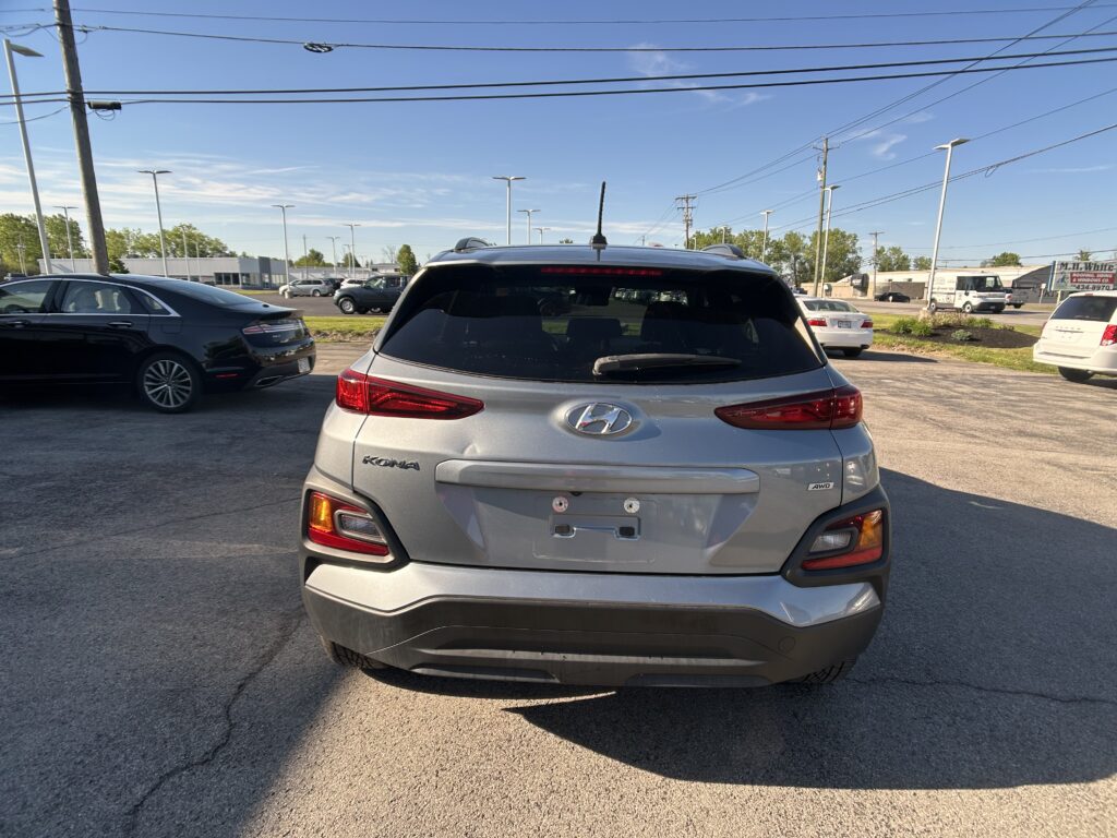 Rear view of a silver Hyundai Kona SUV in a parking lot with other cars and power lines overhead against a clear blue sky.