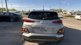 Rear view of a silver Hyundai Kona SUV in a parking lot with other cars and power lines overhead against a clear blue sky.