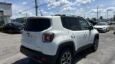 White Jeep Renegade SUV parked in a dealership lot with other cars, under a bright blue sky and power lines overhead.