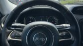 Front view from the driver seat showing the Jeep steering wheel and dashboard, with a parking lot and blue sky with clouds outside through the windshield.