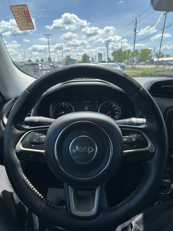 Front view from the driver seat showing the Jeep steering wheel and dashboard, with a parking lot and blue sky with clouds outside through the windshield.