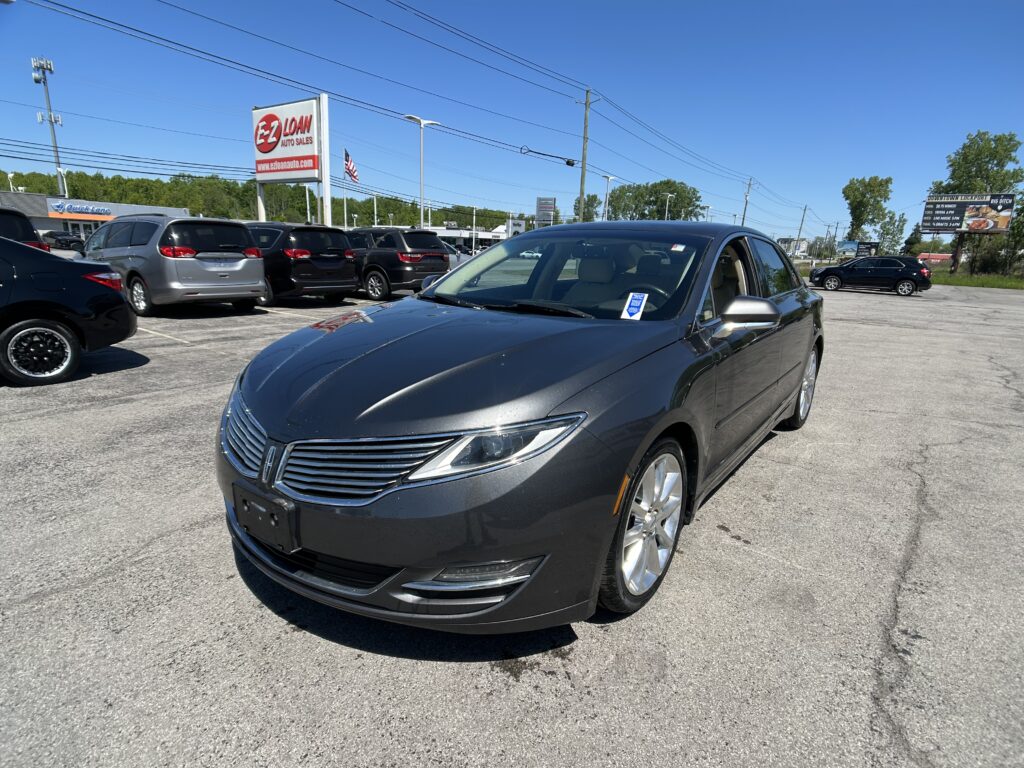 Close-up of a gray sedan in the foreground at a busy car dealership lot, with multiple parked cars, an EZ LOAN sign, and clear blue sky in the background.