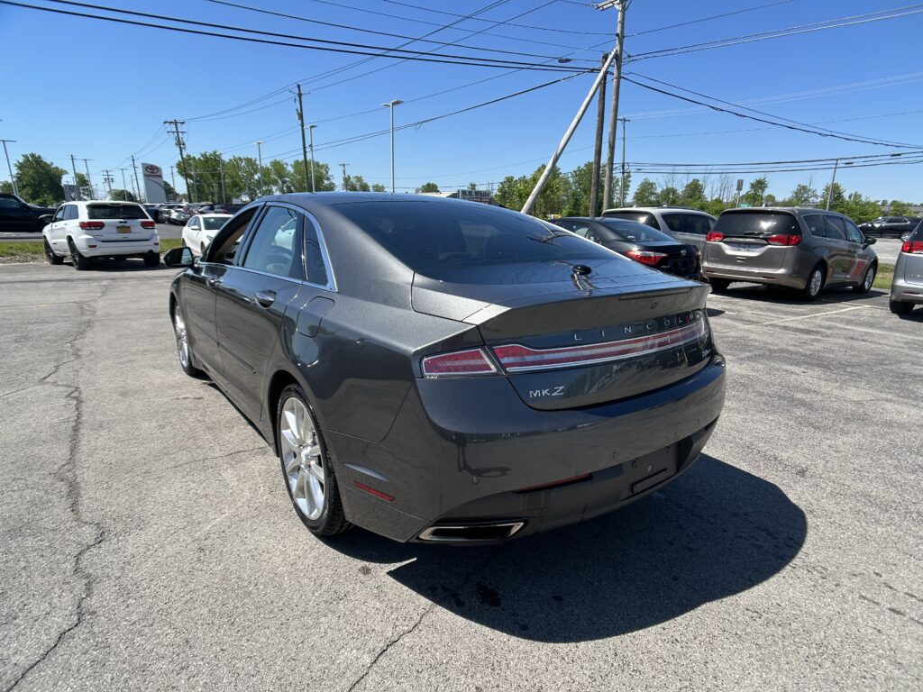 Rear three-quarter view of a gray Lincoln MKZ parked in a busy car lot with other vehicles and power lines overhead under a clear blue sky.