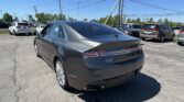 Rear three-quarter view of a gray Lincoln MKZ parked in a busy car lot with other vehicles and power lines overhead under a clear blue sky.