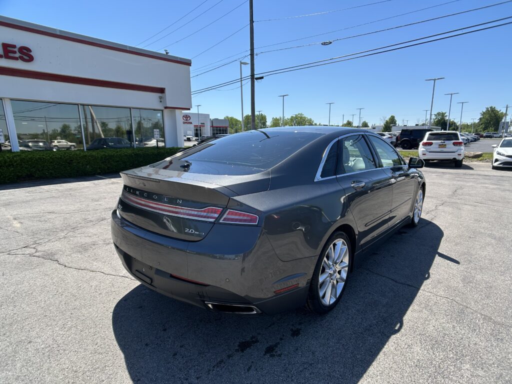 Rear three-quarter view of a dark Lincoln sedan in a car dealership parking lot, with a glass-front showroom on the left and other cars in the background on a sunny day.