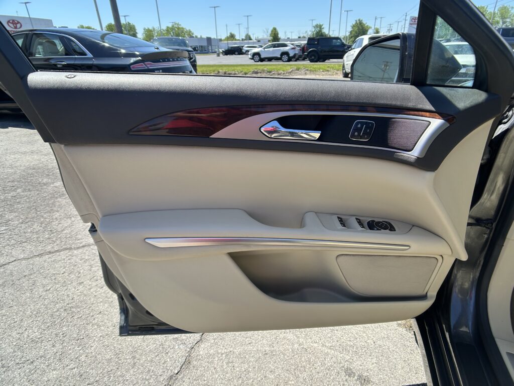 Close-up of a beige car door interior: black upper panel, wooden trim, chrome handle, and power window controls; outside is a car dealership lot.