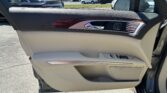 Close-up of a beige car door interior: black upper panel, wooden trim, chrome handle, and power window controls; outside is a car dealership lot.