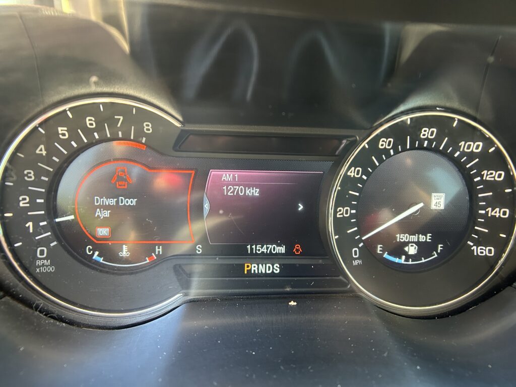 Car dashboard with tachometer on left, speedometer on right, central warning says 'Driver Door Ajar' and odometer at 115470 miles.