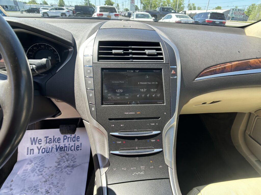 Interior of a car dashboard and center console with a touchscreen, gear selector rail, and climate controls, viewed from the driver's seat; outside is a dealership lot with cars.
