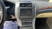 Interior of a car dashboard and center console with a touchscreen, gear selector rail, and climate controls, viewed from the driver's seat; outside is a dealership lot with cars.