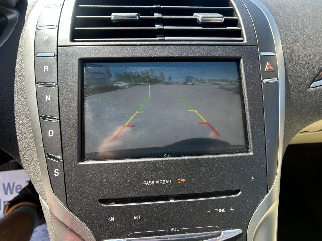 Car infotainment screen showing rearview camera with colored guide lines (green, yellow, red) over a parking lot view
