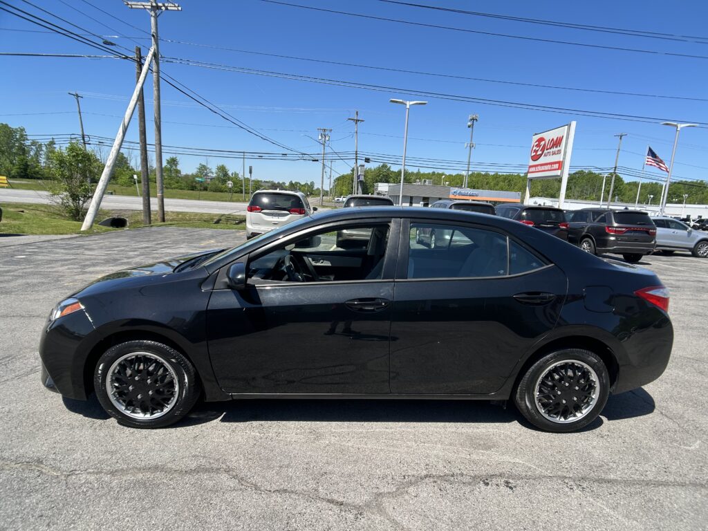 Close-up view of a glossy black sedan with aftermarket wheels parked in a car lot, other vehicles and an EZ Auto Sales sign visible in the background under a blue sky.