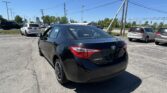 Rear three-quarter view of a black Toyota Corolla in a crowded parking lot with other vehicles and power lines overhead to the blue sky.