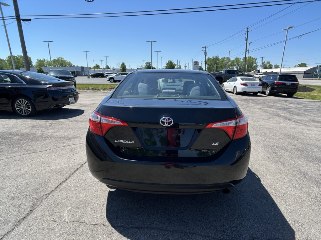 Rear view of a black Toyota Corolla LE in a dealership parking lot, with other cars and a blue sky in the background.