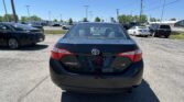 Rear view of a black Toyota Corolla LE in a dealership parking lot, with other cars and a blue sky in the background.