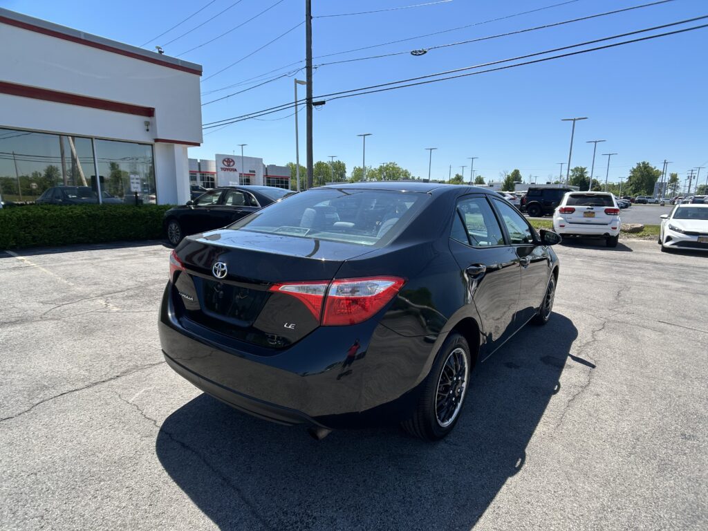 Black Toyota sedan in the foreground parked in a dealership lot with a white building and other cars under a clear blue sky.