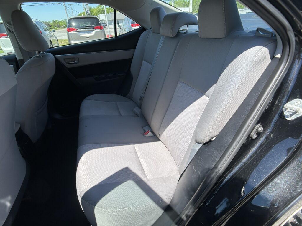 Rear seat interior of a gray fabric sedan with three seats and headrests, seen through an open door while parked outside.
