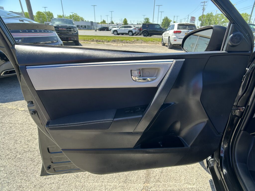 Door panel of a car interior with a chrome handle and beige/black trim; outside a sunny parking lot with cars is visible through the window.