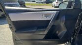 Door panel of a car interior with a chrome handle and beige/black trim; outside a sunny parking lot with cars is visible through the window.