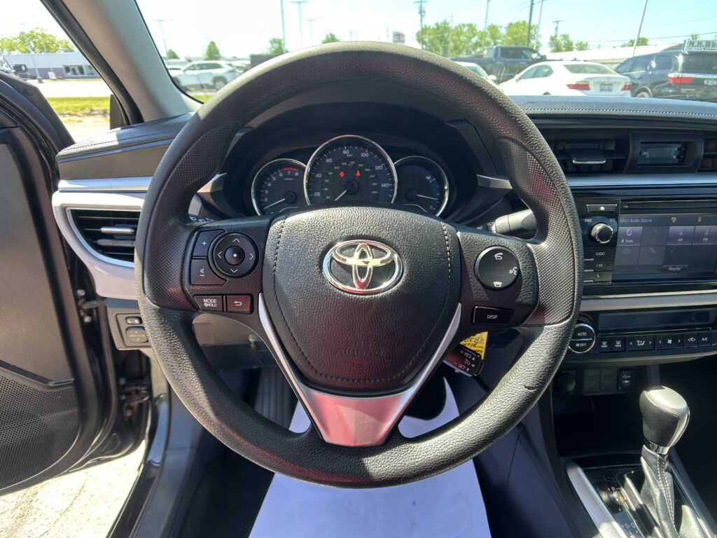Driver’s-seat view of a car interior: steering wheel with Toyota emblem, instrument cluster, and center console; parking lot outside through windshield.