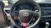 Driver’s-seat view of a car interior: steering wheel with Toyota emblem, instrument cluster, and center console; parking lot outside through windshield.