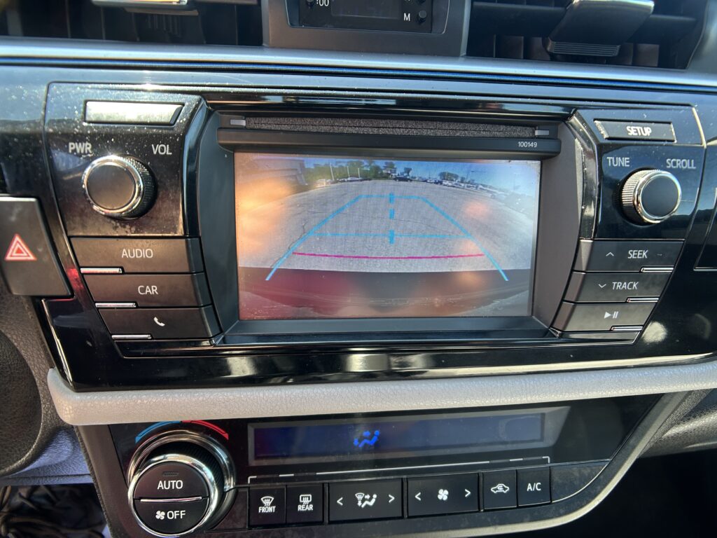 Rearview camera feed on the car's center screen showing blue parking guide lines for backing up, with dashboard controls around it.
