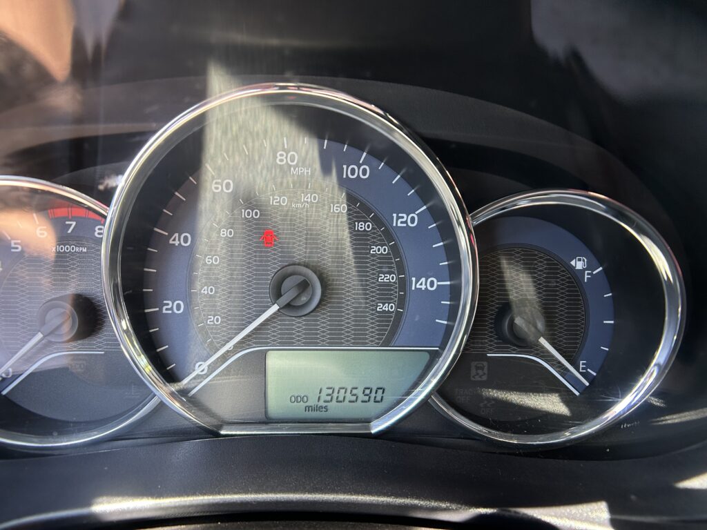Close-up of a car's instrument cluster: speedometer up to 140 mph and odometer reading 130,590 miles with a red warning light visible on the dashboard.