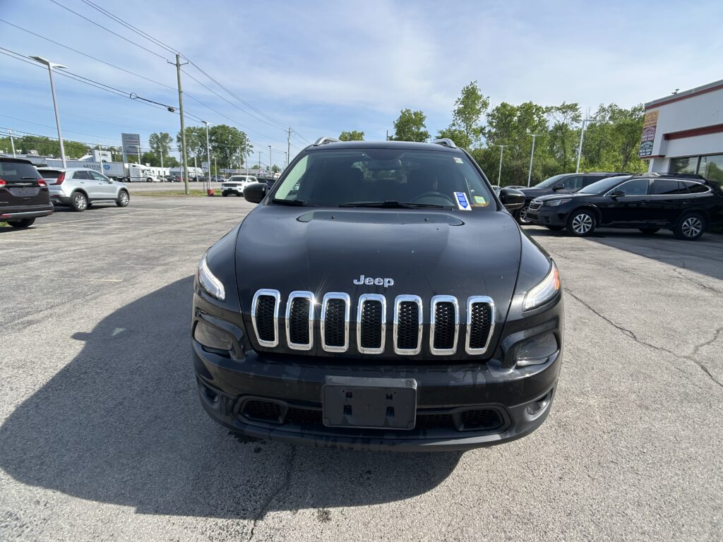 Front view of a black Jeep SUV parked in a dealership lot with other cars and a building in the background.