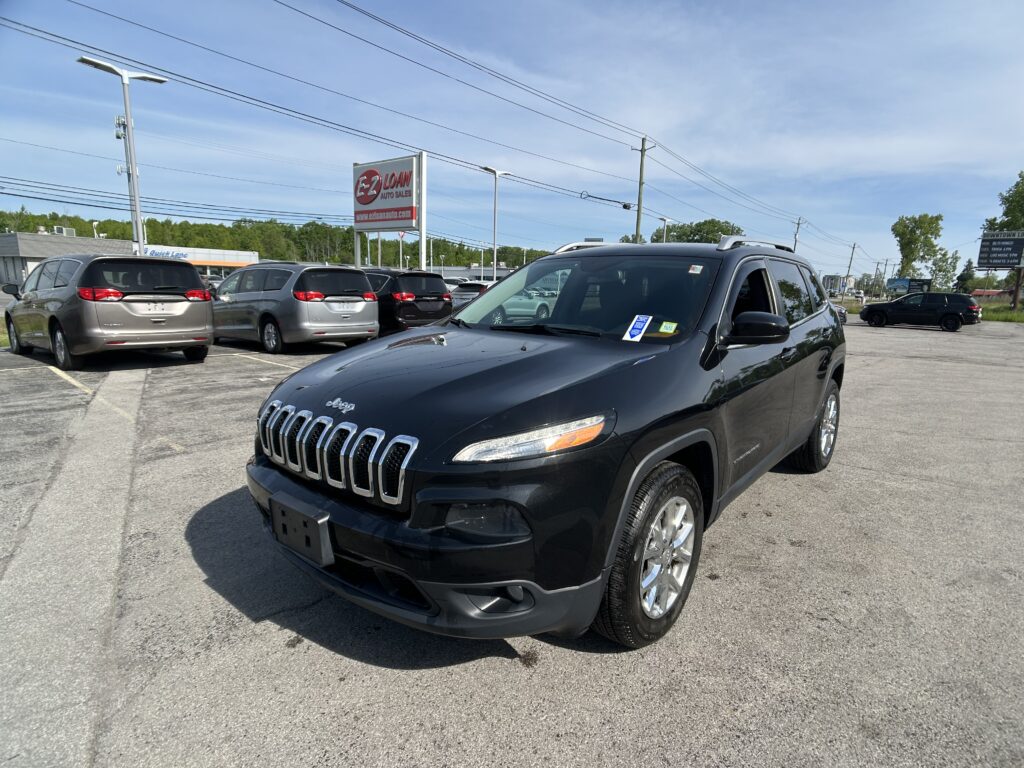 Front-right view of a black Jeep SUV in a car dealership lot, with other minivans parked and an EZ LOAN Auto Sales sign in the background under a blue sky.