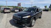 Front-right view of a black Jeep SUV in a car dealership lot, with other minivans parked and an EZ LOAN Auto Sales sign in the background under a blue sky.