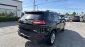 Black Jeep SUV in a dealership lot, rear and side view with other cars and a white building in the background under a blue sky.