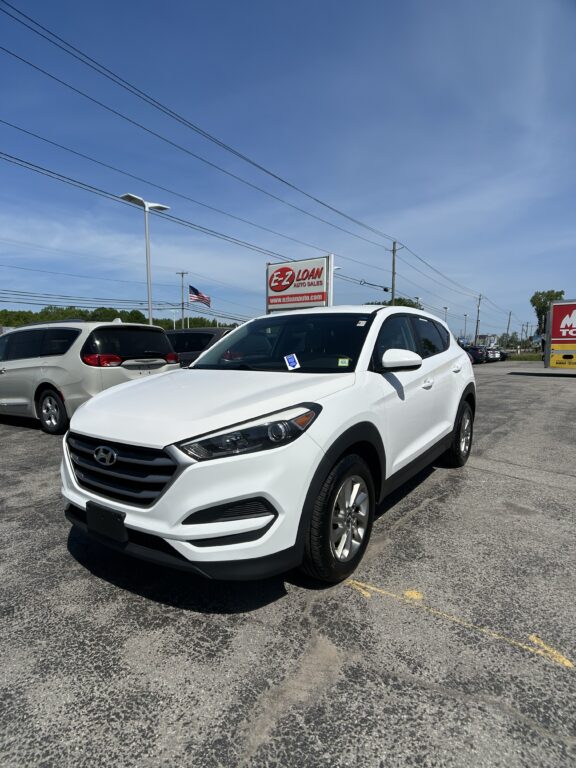 White Hyundai SUV in a car dealership lot with a red EZ LOAN Auto Sales sign in the background and an American flag nearby.