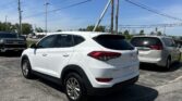 White Hyundai Tucson SUV parked in a lot with another car nearby under a blue sky and overhead power lines.