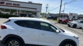 White SUV in a dealership lot, with a building that reads 'AUTO SALES' and other vehicles visible in the background under a clear blue sky.