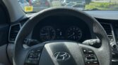 Front-seat view of a car: steering wheel and dashboard, with a dealership lot full of cars outside on a sunny day.