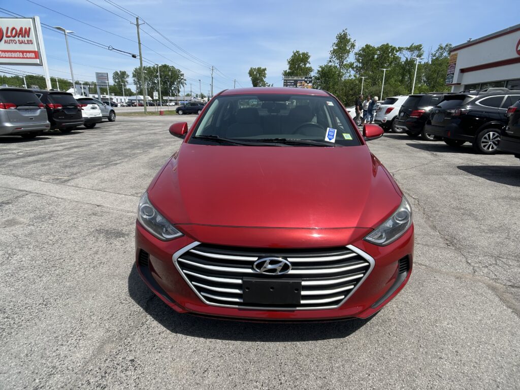 Front view of a red Hyundai in a busy dealership parking lot with other cars and a sign visible in the background.