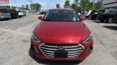 Front view of a red Hyundai in a busy dealership parking lot with other cars and a sign visible in the background.