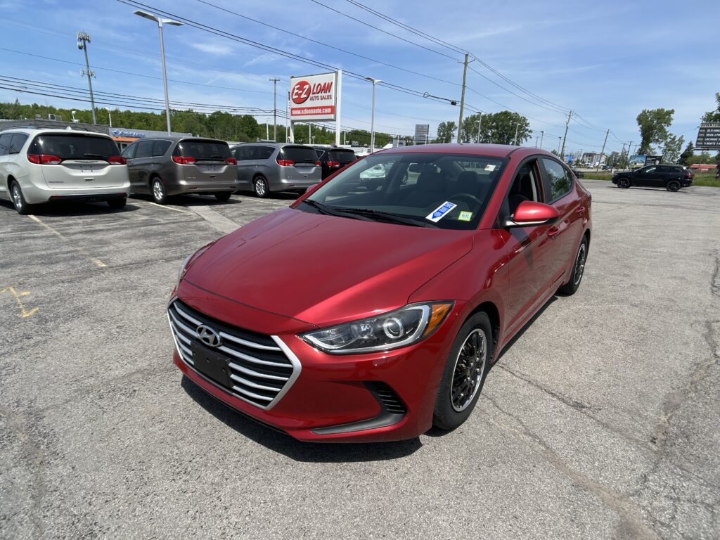 Red compact sedan in the foreground at a car dealership lot, with several gray minivans lined up behind it and a tall EZ LOAN sign in the distance.