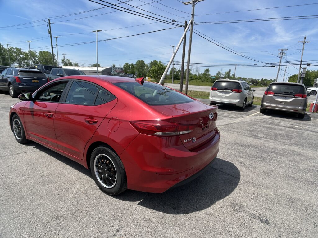 Rear three-quarter view of a red Hyundai Elantra in a parking lot, with other cars and utility poles against a blue sky.