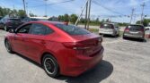 Rear three-quarter view of a red Hyundai Elantra in a parking lot, with other cars and utility poles against a blue sky.
