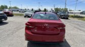 Rear view of a red Hyundai Elantra SE in a parking lot, with more cars and a dealership in the background under a blue sky.
