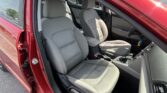 Open-car-door view of a red sedan's gray cloth front seats, center console, and steering wheel with a parking lot visible outside.