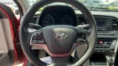 Driver's viewpoint inside a car with steering wheel at center, dashboard gauges visible, and center console radio controls on the right; outside the windshield are parked cars and a bright day.